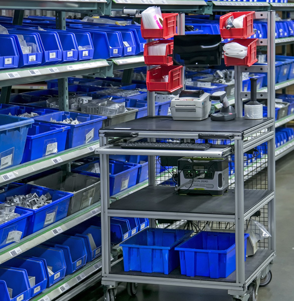 Mobile T-slot aluminum material handling workstation with barcode scanner, label printer, monitor, storage bins, and keyboard tray used for warehouse order fulfillment and inventory management.