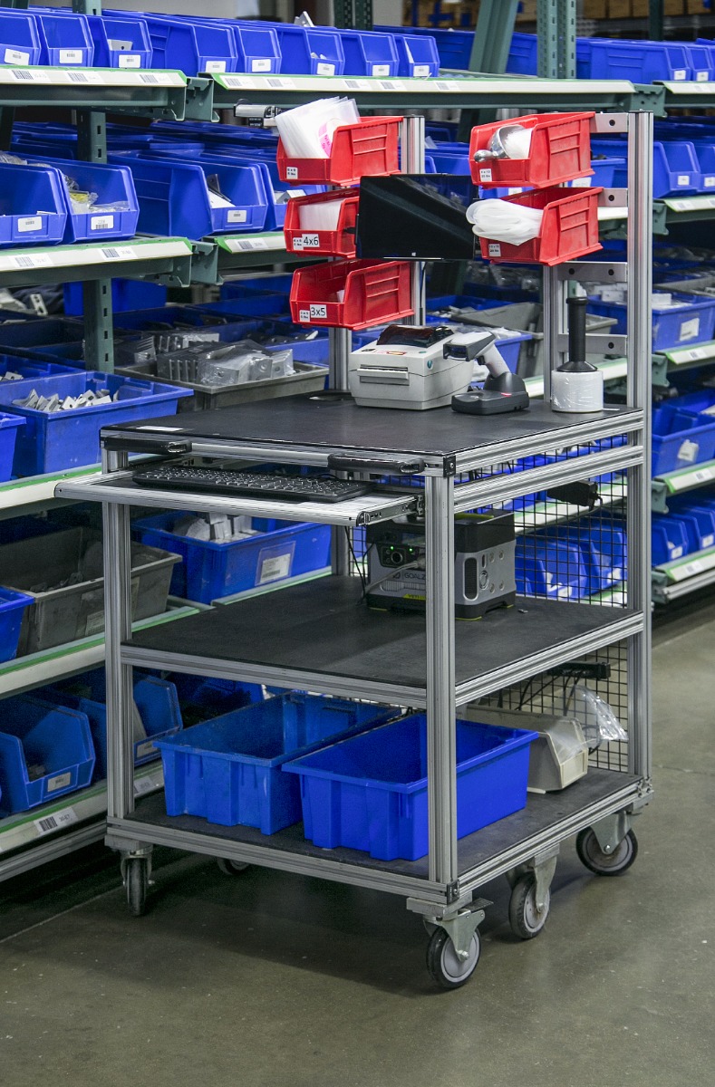 80/20 mobile T-slot aluminum kitting cart with integrated monitor, barcode scanner, keyboard tray, storage bins, wire mesh panel, and shelves for warehouse order picking and lean material handling operations.