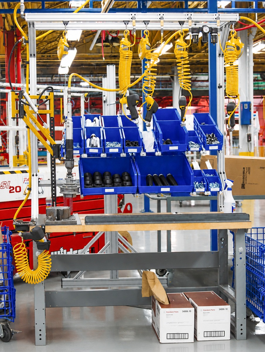 Automotive assembly workstation featuring organized parts bins, suspended pneumatic tools, aluminum extrusion framing, and ergonomic workbench design for lean manufacturing and efficient production.