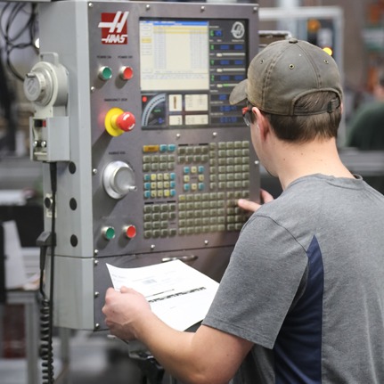 A dedicated fabrication specialist operating a Haas CNC machine control panel. The technician is reviewing technical project specifications on a printed document while preparing to machine custom 80/20 T-slot aluminum components.
