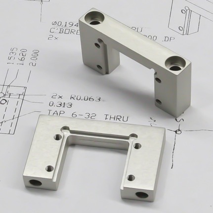 Two custom-machined silver aluminum brackets resting on a technical engineering drawing. The blueprint shows detailed tap and bore specifications, highlighting the high-tolerance fabrication and quality control for 80/20 T-slot components.