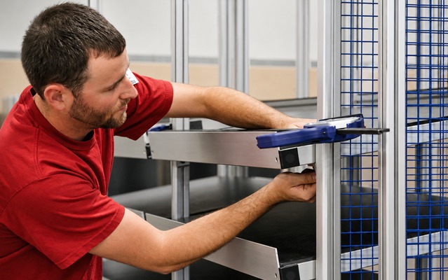 80/20 technician assembling a custom T-slot aluminum structure with shelving and wire mesh panels, demonstrating precision assembly, quality craftsmanship, and custom industrial build services.