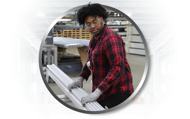 A warehouse specialist wearing safety glasses and gloves carefully handling a bundle of silver 80/20 T-slot aluminum extrusions. The specialist is preparing the material for shipment in a professional industrial fulfillment center.