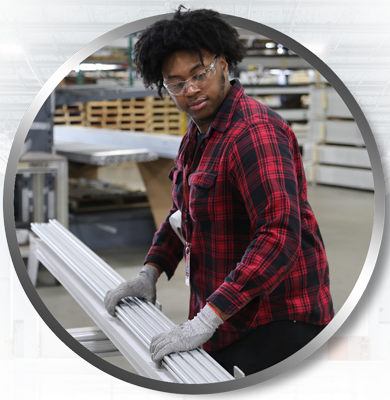 A warehouse specialist wearing safety glasses and gloves carefully handling a bundle of silver 80/20 T-slot aluminum extrusions. The specialist is preparing the material for shipment in a professional industrial fulfillment center.