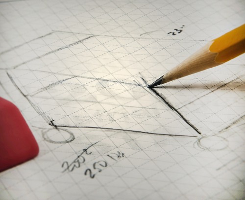 Close-up of a pencil sketching a technical design for a mobile cart on graph paper. The hand-drawn concept includes dimensions and notes, representing the early planning stage of a custom 80/20 T-slot aluminum assembly.