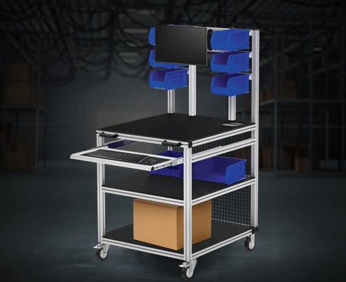 A finished industrial mobile workstation made from 80/20 T-slot aluminum framing. The unit features a black tabletop, integrated blue storage bins, a slide-out keyboard tray with a computer monitor mount, and heavy-duty casters, shown in a warehouse.