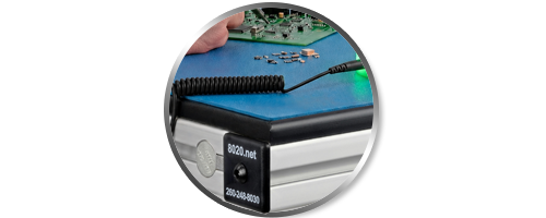 Close-up circular inset of an ESD-safe 80/20 workstation featuring a blue dissipative mat, a coiled grounding wrist strap cord, and a snap connector. An operator is shown working on a circuit board in the background, highlighting static-control measures i
