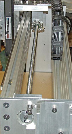 Close-up of a mechanical linear motion system with metal profiles, lead screw, and cable carrier, possibly part of a CNC machine setup.