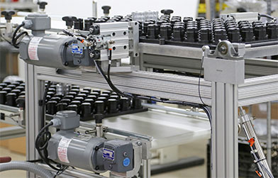 An industrial conveyor system with multiple electric motors and pneumatic actuators, handling a series of black cylindrical objects.