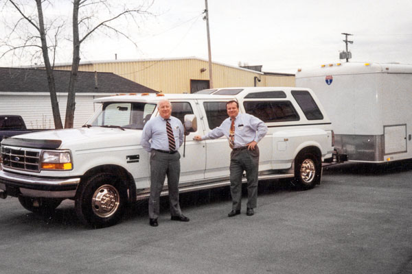 Two men in business attire standing next to a white pickup truck with a trailer in the background.