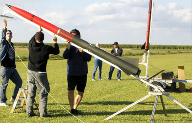 Individuals setting up a model rocket on a launch pad in an open field with clear skies.
