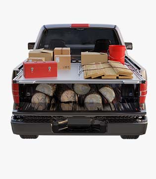 Pickup truck bed filled with an organized storage system of shelves holding tools, boxes, and equipment, with wood logs underneath.